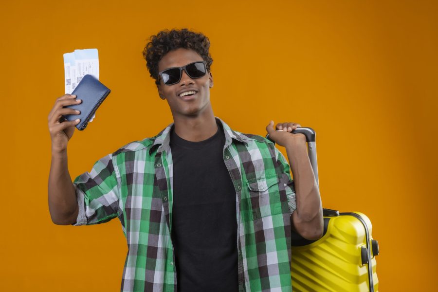 young african american traveler man wearing black sunglasses standing with suitcase holding air tickets smiling cheerfully positive and happy over orange background